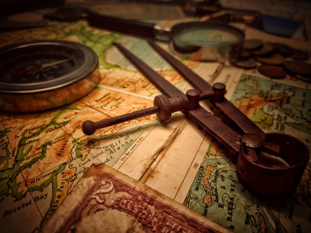 A vintage setup consisting of a map, an old-fashioned compass, a pair of dividers, and some coins. The lighting gives a warm, nostalgic feel. The map appears detailed, showing various geographic features and regions. There is also a partial view of a vintage currency note with intricate designs.