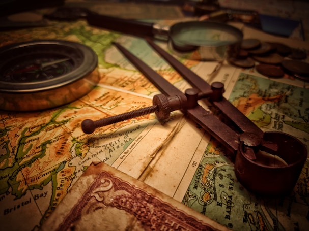 A vintage setup consisting of a map, an old-fashioned compass, a pair of dividers, and some coins. The lighting gives a warm, nostalgic feel. The map appears detailed, showing various geographic features and regions. There is also a partial view of a vintage currency note with intricate designs.