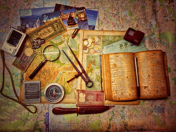 A vintage travel-themed collection of items is arranged on an old map. Coins, banknotes, and postcards are scattered alongside an open book with yellowed pages. A magnifying glass and a compass suggest exploration. Other objects include a camera, a map, and writing implements.