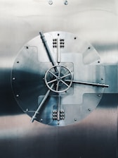 A large, round metal vault door with intricate handles and mechanisms, prominently featuring a central wheel with spokes. The surface is smooth and metallic, reflecting a subdued light.