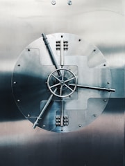 A large, round metal vault door with intricate handles and mechanisms, prominently featuring a central wheel with spokes. The surface is smooth and metallic, reflecting a subdued light.