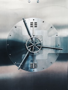A large, round metal vault door with intricate handles and mechanisms, prominently featuring a central wheel with spokes. The surface is smooth and metallic, reflecting a subdued light.