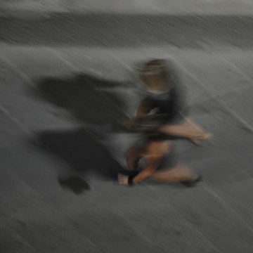 A blurred image capturing the motion of a person, casting a prominent shadow on a textured grey surface. The figure is in mid-movement, creating a dynamic and abstract appearance.