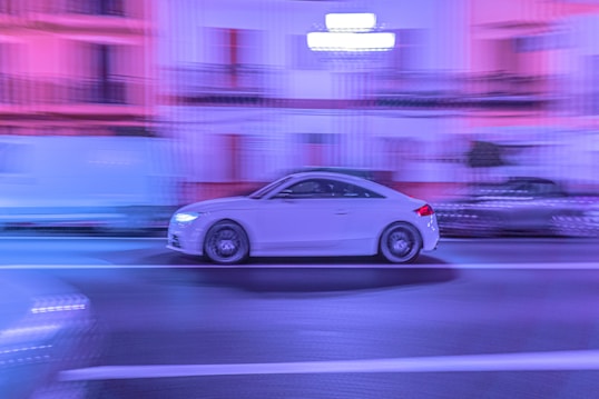 A white sports car is captured moving rapidly on a street, with a blurred background of buildings illuminated in purple and blue lights. The photo creates a sense of speed and dynamism through motion blur.