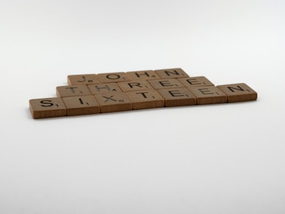Wooden letter tiles are arranged on a flat surface to form the words 'JOHN THREE SIXTEEN'. The tiles appear to be from a board game, similar to Scrabble.