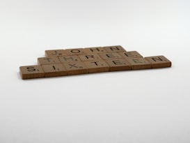 Wooden letter tiles are arranged on a flat surface to form the words 'JOHN THREE SIXTEEN'. The tiles appear to be from a board game, similar to Scrabble.
