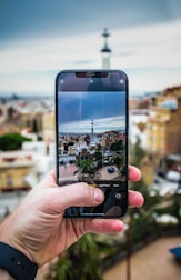 A hand holding a smartphone capturing a cityscape. The smartphone screen displays a detailed view of a city with various buildings and a prominent tower in the center. The background is blurred, showcasing a panoramic view of the city with blue sky and some clouds.