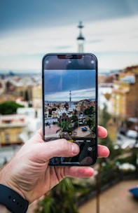 A hand holding a smartphone capturing a cityscape. The smartphone screen displays a detailed view of a city with various buildings and a prominent tower in the center. The background is blurred, showcasing a panoramic view of the city with blue sky and some clouds.