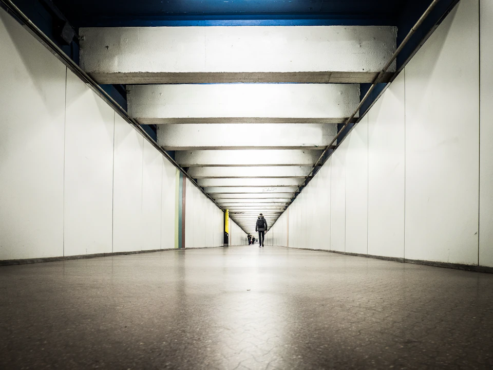 Long, narrow tunnel with white walls and blue beams, illuminated by a single overhead light