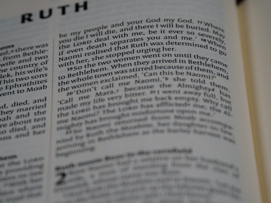 A close-up view of a page from a Bible showing text from the Book of Ruth. The text is in English and printed in a clear font. The pages appear slightly yellowed, indicating the book may be older or well-used. The lighting is soft, creating shadows along the folds of the page.