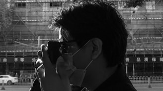 A person wearing glasses and a face mask is holding a camera up to their face, seemingly taking a photo. The background features scaffolding on a building, along with some signage with Chinese characters. The image is in black and white, highlighting shadows and contrast.