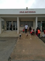 A group of people is gathered outside a building with the sign 'VOLS NATIONAUX.' The area in front of the building has a ramp where two small goats are walking. Some people are sitting on the steps, and a couple of individuals are wearing orange vests. The atmosphere appears casual, and the area is well-lit.