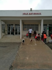 A group of people is gathered outside a building with the sign 'VOLS NATIONAUX.' The area in front of the building has a ramp where two small goats are walking. Some people are sitting on the steps, and a couple of individuals are wearing orange vests. The atmosphere appears casual, and the area is well-lit.