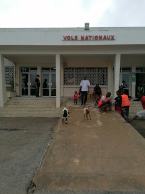 A group of people is gathered outside a building with the sign 'VOLS NATIONAUX.' The area in front of the building has a ramp where two small goats are walking. Some people are sitting on the steps, and a couple of individuals are wearing orange vests. The atmosphere appears casual, and the area is well-lit.