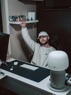 A man wearing a white cap and sweatshirt is sitting at a desk with a computer monitor, keyboard, and mouse. He is holding up a peace sign with his right hand. The desk is also equipped with a smart speaker and a remote control. Behind him, a shelf displays several figurines and cards.