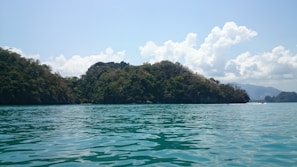 A serene seascape with calm turquoise waters and lush green islands under a bright blue sky. Puffy white clouds float above the islands, adding depth to the horizon. The tranquility of the scene is enhanced by the soft ripples on the water's surface.