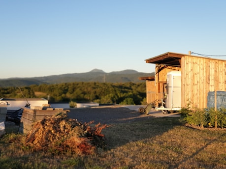 A wooden shed sits in a rural area with visible mountains in the background under a clear blue sky. There is a pile of dried plants and some gardening tools nearby.