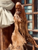 A wooden sculpture of a bearded figure draped in flowing garments. The detailed carving highlights the figure's facial expressions and textured clothing. Sunlight enhances the natural wood grain, casting shadows that add depth. A soft, light-colored fabric is draped in the background, and wooden window frames are visible.