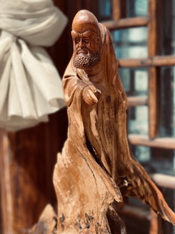 A wooden sculpture of a bearded figure draped in flowing garments. The detailed carving highlights the figure's facial expressions and textured clothing. Sunlight enhances the natural wood grain, casting shadows that add depth. A soft, light-colored fabric is draped in the background, and wooden window frames are visible.