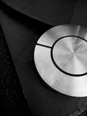 A circular metallic object with a brushed texture is positioned on a dark, geometric, textured background. The metallic object features concentric circles and segments, creating a sense of symmetry and design sophistication.
