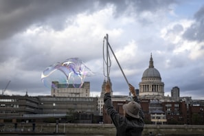 A person wearing a hat is performing a bubble trick using a long stick, with a large, iridescent bubble floating in the air. The background features a cityscape with a prominent dome structure under a cloudy sky.