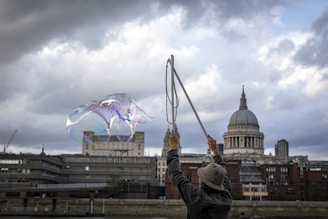 A person wearing a hat is performing a bubble trick using a long stick, with a large, iridescent bubble floating in the air. The background features a cityscape with a prominent dome structure under a cloudy sky.