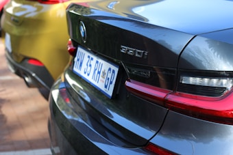 Close-up view of the rear ends of two cars parked closely together. One car is a shiny dark gray BMW 330i with visible taillights and a clear view of the license plate from South Africa. The other car is a metallic gold or yellow color.
