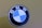 A close-up view of a BMW logo, featuring alternating blue and white quadrants in a circular emblem with the letters B, M, and W at the top. The background is a muted olive green color.