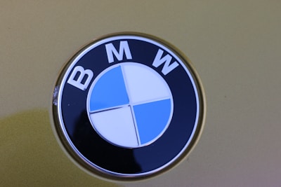 A close-up view of a BMW logo, featuring alternating blue and white quadrants in a circular emblem with the letters B, M, and W at the top. The background is a muted olive green color.
