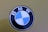 A close-up view of a BMW logo, featuring alternating blue and white quadrants in a circular emblem with the letters B, M, and W at the top. The background is a muted olive green color.