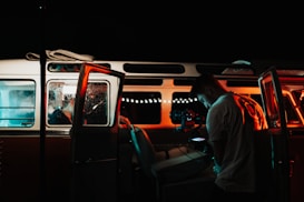 A person is standing outside a vintage minivan, illuminated by blue and red lights. They are operating a camera setup, capturing something inside the vehicle. The interior is dimly lit, suggesting a focus on nighttime or low-light conditions. The scene has a cinematic quality with strong color contrasts from the lighting.