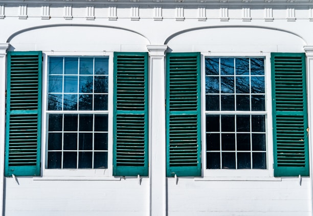 Two large windows with multiple panes, each flanked by green shutters on a white building. The windows reflect the blue sky and tree branches, suggesting an outdoor scene.