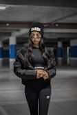 A person is standing in a dimly lit parking garage, wearing a black beanie with the word 'Queen' and sunglasses. Dressed in a black puffer jacket and matching athletic wear, the individual exudes a sense of confidence and style.