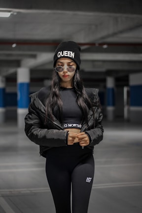 A person is standing in a dimly lit parking garage, wearing a black beanie with the word 'Queen' and sunglasses. Dressed in a black puffer jacket and matching athletic wear, the individual exudes a sense of confidence and style.