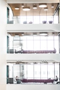 A modern, multi-level interior space with glass railings, wood-paneled ceilings, and large windows. Each level features seating areas with purple couches and a few people sitting, engaged in work or conversation. The natural light filters through the windows, creating a bright and airy atmosphere.