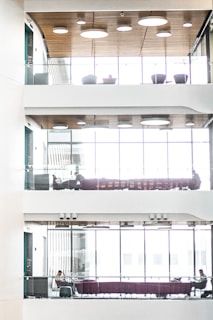 A modern, multi-level interior space with glass railings, wood-paneled ceilings, and large windows. Each level features seating areas with purple couches and a few people sitting, engaged in work or conversation. The natural light filters through the windows, creating a bright and airy atmosphere.