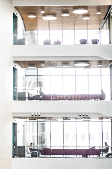 A modern, multi-level interior space with glass railings, wood-paneled ceilings, and large windows. Each level features seating areas with purple couches and a few people sitting, engaged in work or conversation. The natural light filters through the windows, creating a bright and airy atmosphere.