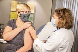 A person wearing a mask receives a vaccination from a healthcare professional, also wearing a mask, in a medical office setting. The professional is dressed in a white coat, indicating a clinical environment. Documented information is visible in the background on the walls.