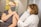A person wearing a mask receives a vaccination from a healthcare professional, also wearing a mask, in a medical office setting. The professional is dressed in a white coat, indicating a clinical environment. Documented information is visible in the background on the walls.