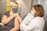 A person wearing a mask receives a vaccination from a healthcare professional, also wearing a mask, in a medical office setting. The professional is dressed in a white coat, indicating a clinical environment. Documented information is visible in the background on the walls.