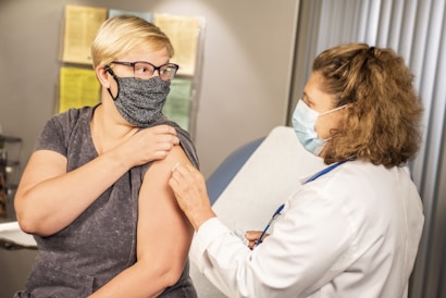 A person wearing a mask receives a vaccination from a healthcare professional, also wearing a mask, in a medical office setting. The professional is dressed in a white coat, indicating a clinical environment. Documented information is visible in the background on the walls.