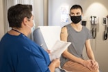 Two people wearing masks are engaged in a conversation in what appears to be a medical setting. One of them, dressed in a blue uniform, is holding a clipboard with papers. The other person is sitting on a medical examination table, wearing a striped shirt with a small adhesive bandage on their upper arm. Medical equipment is mounted on the wall in the background.