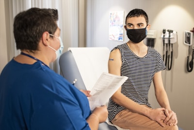 Two people wearing masks are engaged in a conversation in what appears to be a medical setting. One of them, dressed in a blue uniform, is holding a clipboard with papers. The other person is sitting on a medical examination table, wearing a striped shirt with a small adhesive bandage on their upper arm. Medical equipment is mounted on the wall in the background.