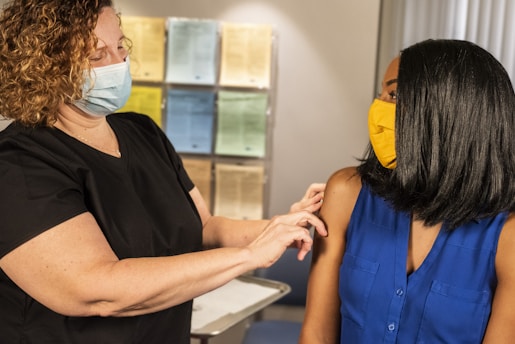 A healthcare professional wearing a light blue face mask is preparing to administer a vaccine to a woman wearing a yellow face mask and a blue sleeveless top. They are in an indoor setting, possibly a clinic or medical office, with documents and papers visible in the background.