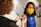 A person with curly hair assists another person who is wearing a yellow face mask and a blue sleeveless shirt. The person assists in placing a small bandage on the other's arm, indicating a medical or vaccination setting. The room contains medical equipment visible in the background, suggesting a clinic environment.