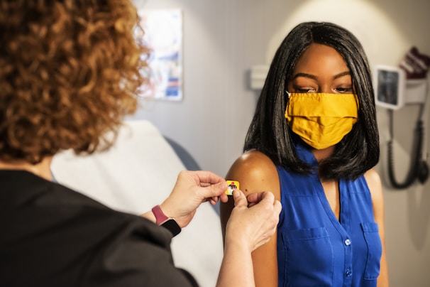 A person with curly hair assists another person who is wearing a yellow face mask and a blue sleeveless shirt. The person assists in placing a small bandage on the other's arm, indicating a medical or vaccination setting. The room contains medical equipment visible in the background, suggesting a clinic environment.