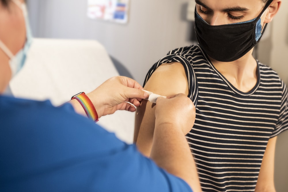 A nurse applies a bandaid to the arm of a thin white patient in a striped shirt. Both are wearing COVID-19 facemasks, the patient wearing two masks. The nurse also wears a rainbow LGBTQ pride bracelet.