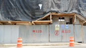 A construction site is enclosed by corrugated metal fencing with warning signs attached. Orange traffic cones are positioned in front of the gate. A black tarp covers the upper part of the site, and the fencing has red numbers painted on it.