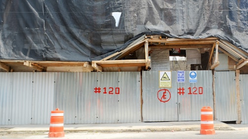 A construction site is enclosed by corrugated metal fencing with warning signs attached. Orange traffic cones are positioned in front of the gate. A black tarp covers the upper part of the site, and the fencing has red numbers painted on it.