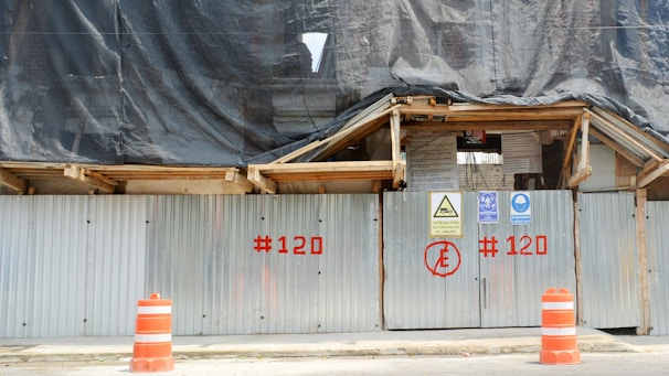 A construction site is enclosed by corrugated metal fencing with warning signs attached. Orange traffic cones are positioned in front of the gate. A black tarp covers the upper part of the site, and the fencing has red numbers painted on it.